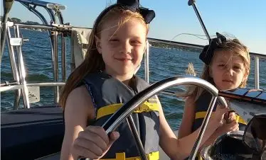 Two girls in life jackets are steering a sailboat on calm waters, with sunlight shining and a clear blue sky in the background.