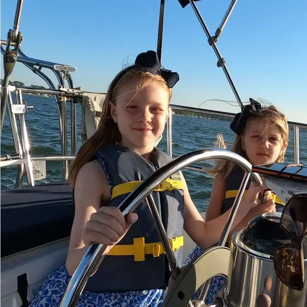 Two girls in life jackets are steering a sailboat on calm waters, with sunlight shining and a clear blue sky in the background.
