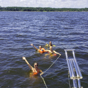 Group of people wearing life jackets in a lake, smiling and waves as they float near a metal ladder with a tree-lined shore in the background.