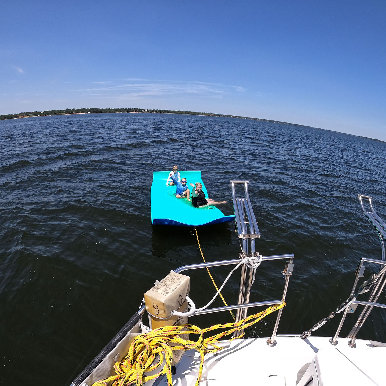 Group of three people riding a bright blue inflatable slip (towed) on a large lake, anchored to a boat with ropes and metal rails visible in the foreground.